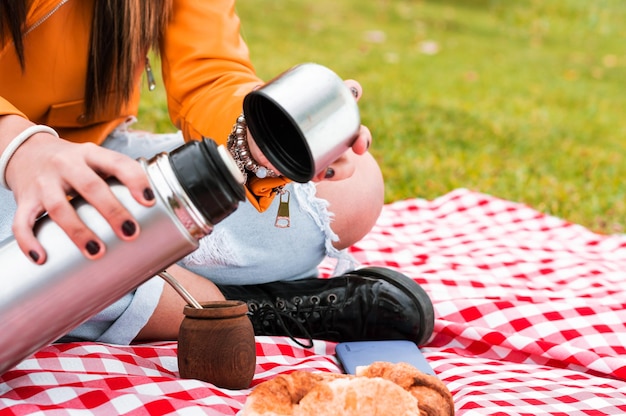 Photo young woman holding a thermos of water sitting in a park with a mate and croissants