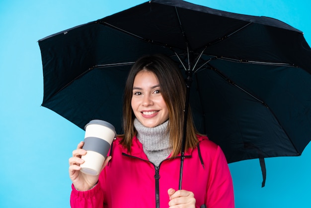 Young woman holding an umbrella and coffee to take away over blue wall