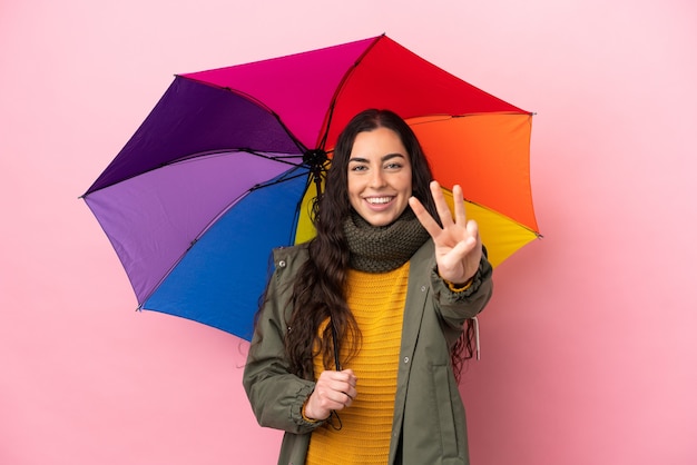 Young woman holding an umbrella isolated on pink background happy and counting three with fingers