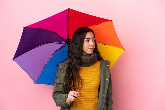 Giovane donna che tiene un ombrello isolato su sfondo rosa guardando di lato