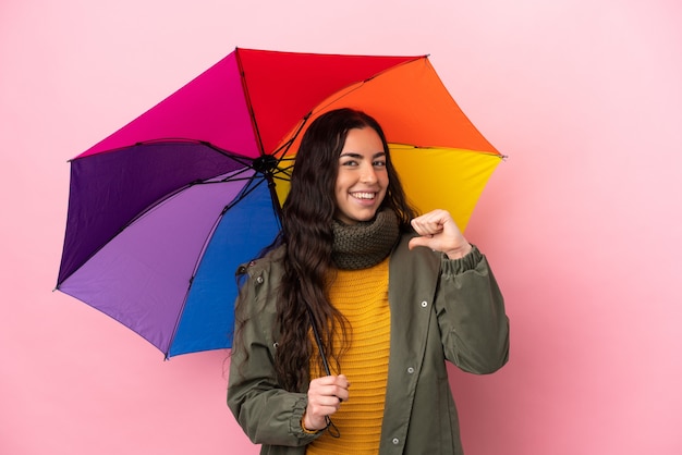 Young woman holding an umbrella isolated on pink background proud and self-satisfied