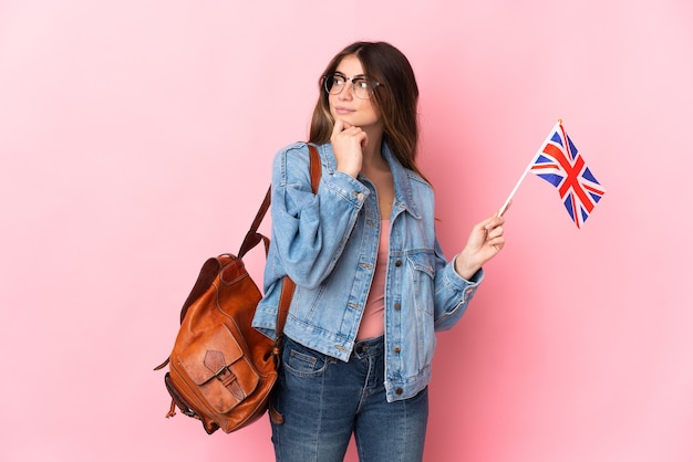 Young woman holding an United Kingdom flag isolated on pink wall and looking up