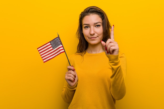 Young woman holding an united states flag showing number one with finger.