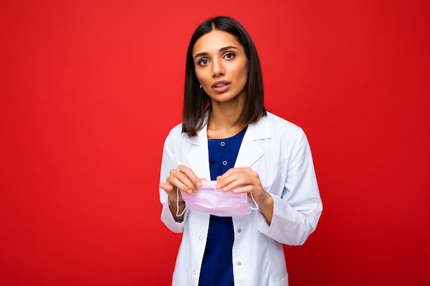 Young Woman holding virus protective mask on face against coronavirus and white medical coat isolated on red background. Free space