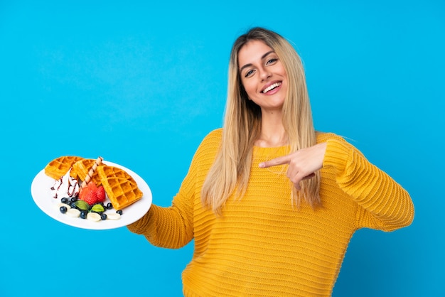 Young woman holding waffles over isolated wall and pointing it