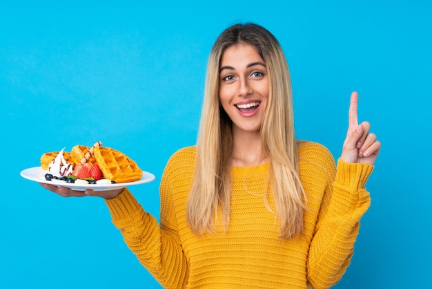 Young woman holding waffles over isolated wall pointing up a great idea