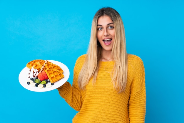 Young woman holding waffles over isolated wall  with surprise and shocked facial expression