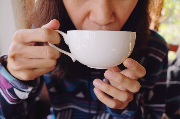 Una giovane donna con una tazza di caffè bianca nelle sue mani e bevendolo al mattino