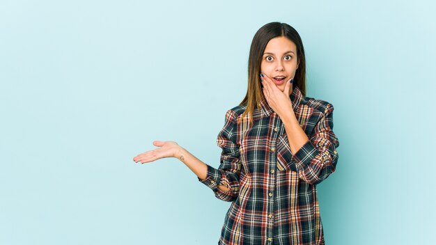 Young woman holds copy space on a palm, keep hand over cheek. Amazed and delighted.