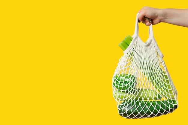 Young woman holds an eco string shopping bag with green vegetables on yellow background