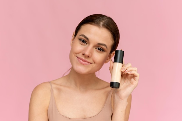 A young woman holds a foundation in a tube on a pink background Makeup advertising