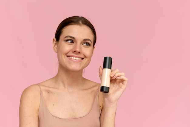 A young woman holds a foundation in a tube on a pink background. Makeup advertising