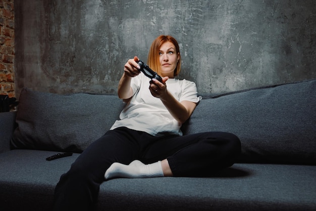 A young woman holds a game joystick while playing video games while sitting on the couch at home