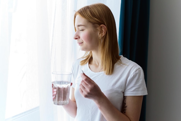 한 젊은 여성이 머리나 복부에 통증을 느끼기 위해 물 한 컵과 알약을 들고 있다
