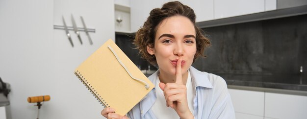 Young woman holds her diary shows secret hush gesture presses finger to lips puts her thoughts in