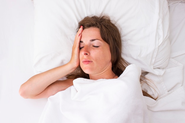 A young woman holds her head and lies in bed. Concept insomnia, headache, malaise, illness. Flat lay, top view