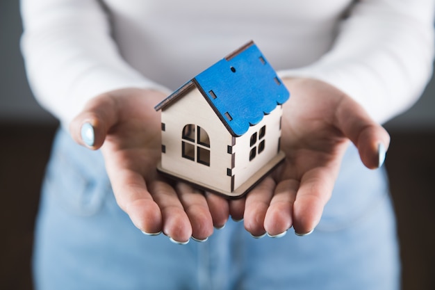 Young woman holds a house in her hands on a gray wall