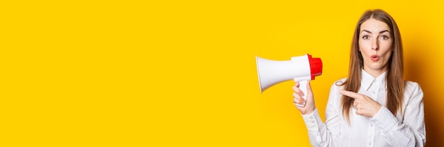 Young woman holds a megaphone in her hands and points her finger at it on a yellow background