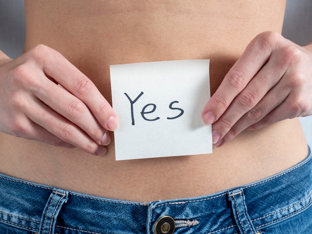 A young woman holds a paper sticker in front of her bare stomach. the piece of paper says Yes