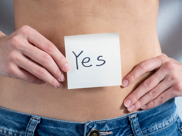A young woman holds a paper sticker in front of her bare stomach which writes yes