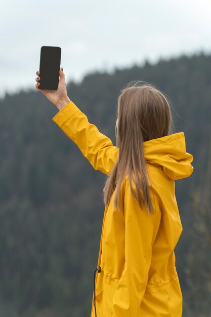 Young woman holds phone in hand with blank empty black screen in yellow jacket in mountains Copy space Mock up