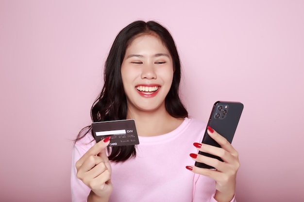 Young woman holds a smartphone and a credit card in her hand Asian woman poses with equipment on light pink background