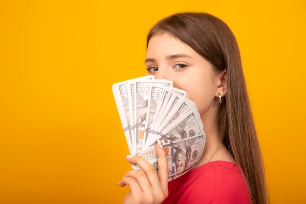 Young woman holds a stack of dollar bills fanned near her face against a bright yellow orange background copy space