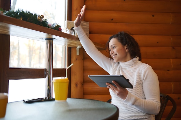 A young woman holds a tablet and waves her hand through the window sitting at the table of a cozy wooden cafe