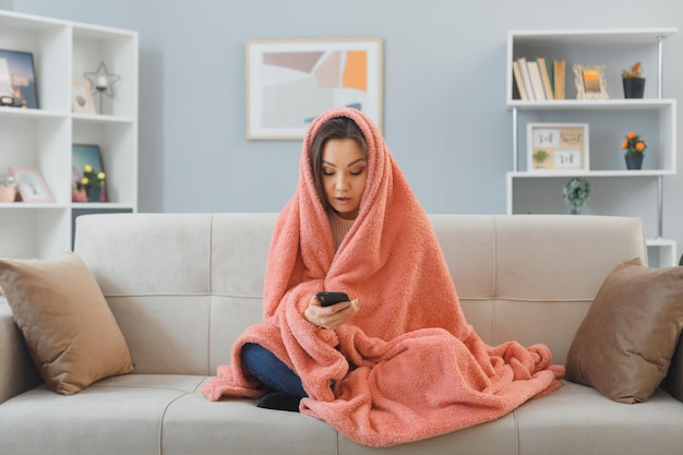 Young woman in home clothes sitting on a couch under blanket texting a message using smartphone looking surprised spending time at home