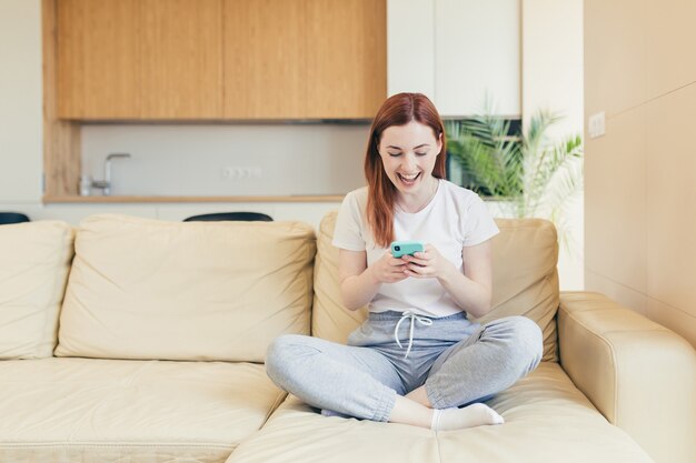 Giovane donna a casa che riceve emozioni positive vincenti e buone notizie mentre legge il telefono mentre è seduto sul divano