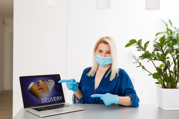 Young woman at home in self-quarantine order products online delivery. Quarantined work from home. Masked womany new life with coronavirus isolation. Remote working on a computer. pandemic lifestyle
