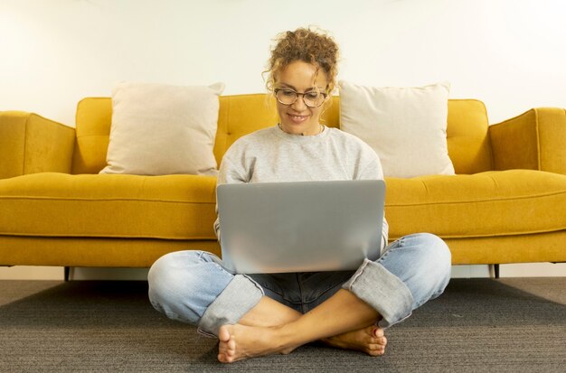 Young woman at home sitting on floor with crossed legs working using laptop in the living room at home. Woman sitting on living room floor working on laptop at home office