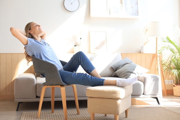 Young woman at home sitting on modern chair in front of window, relaxing in her living room.