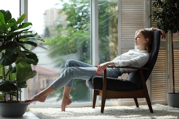 Young woman at home sitting on modern chair near window relaxing in living room