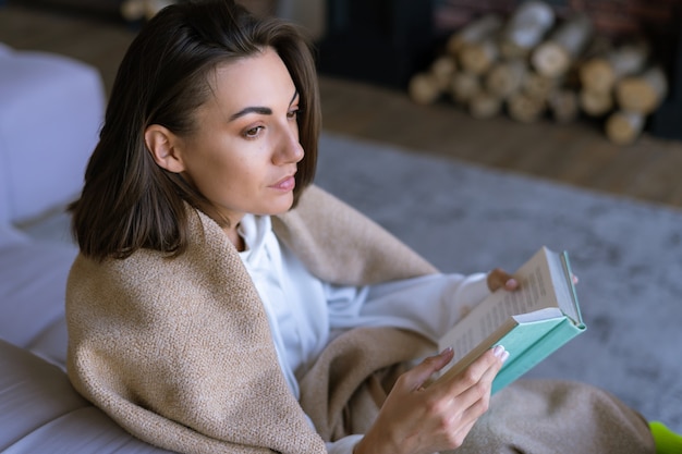 A young woman at home in a white hoodie on the couch wrapped herself in a warm blanket, reads a book