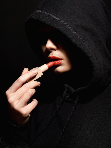 Young woman in hood painting her lips with red lipstick