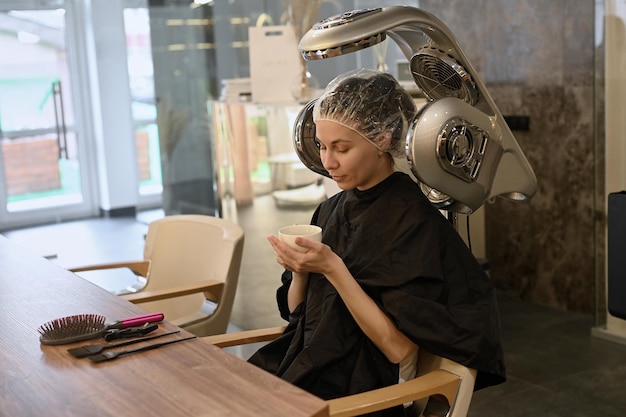 ヘアサロンでフード付きドライヤーマシンの下で若い女性