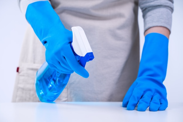 Young woman housekeeper is doing cleaning white table in apron with blue gloves, spray cleaner, wet yellow rag, close up, copy space, blank design concept.