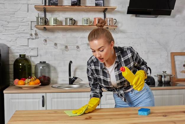 Young woman or housewife in rubber gloves wiping table with microfiber cloth at home kitchen.