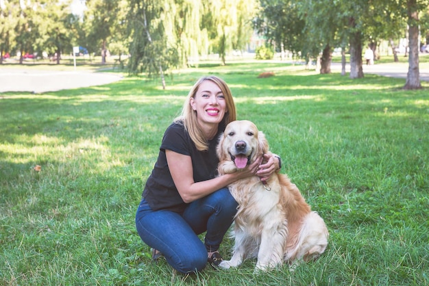 Young woman hugs her retriever while sitting on the grass in a summer park.