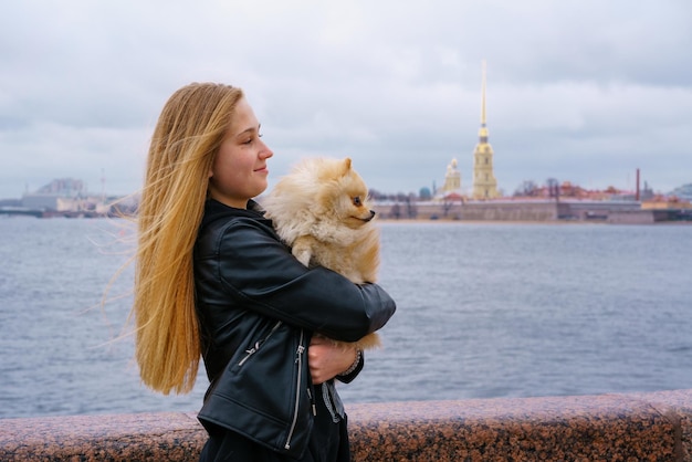 La giovane donna abbraccia il cane dello spitz all'aperto sull'argine del fiume della città in vestiti neri con i capelli lunghi happ...