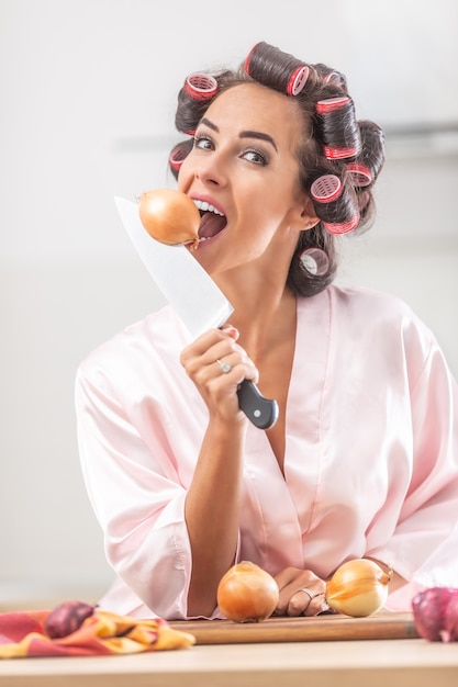 Young woman imitates onion biting with a knife in her hand and curlers in her hair.