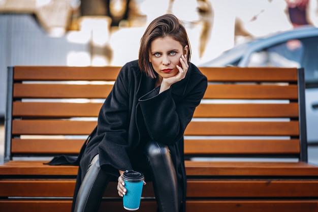 Фото Молодая женщина в черном пальто, сидит на скамейке в городе и держит чашку кофе.