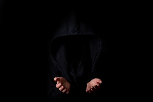 Фото Молодая женщина в черном капюшоне лицо не видно с вытянутыми ладонями на темно-черном фоне.