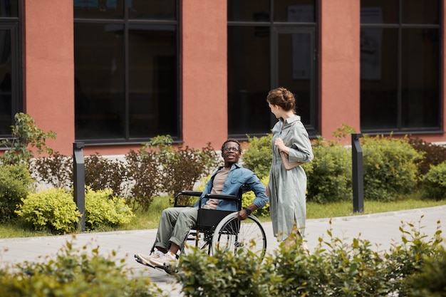 Фото Молодая женщина в платье гуляет по улице и болтает с афро-американским парнем-инвалидом в инвалидной коляске