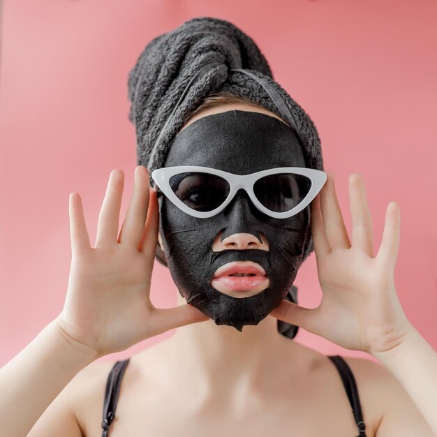 Фото Молодая женщина в очках, использующая черную косметическую ткань для маски лица на розовом фоне.
