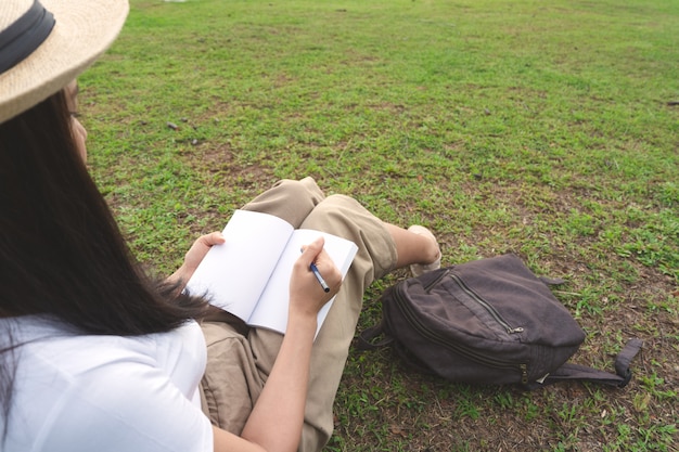 写真 屋外の公園の草の上に鉛筆でノートブックを書く帽子の若い女性。