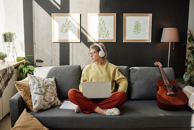 Фото Молодая женщина в наушниках сидит на диване в комнате и работает онлайн на ноутбуке