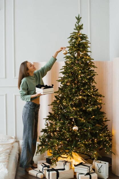 Фото Молодая женщина в домашней одежде украшает рождественскую елку игрушками в уютном доме