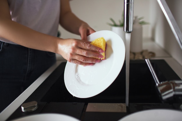 Фото Молодая женщина на кухне во время карантина. белые тарелки с губкой и посудомоечной машиной. уборка посуды в одиночку на кухне. вырезать вид.